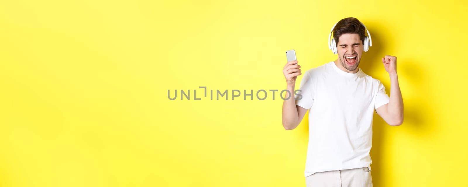 Happy man dancing and listening music in headphones, holding mobile cell phone, standing against yellow background.