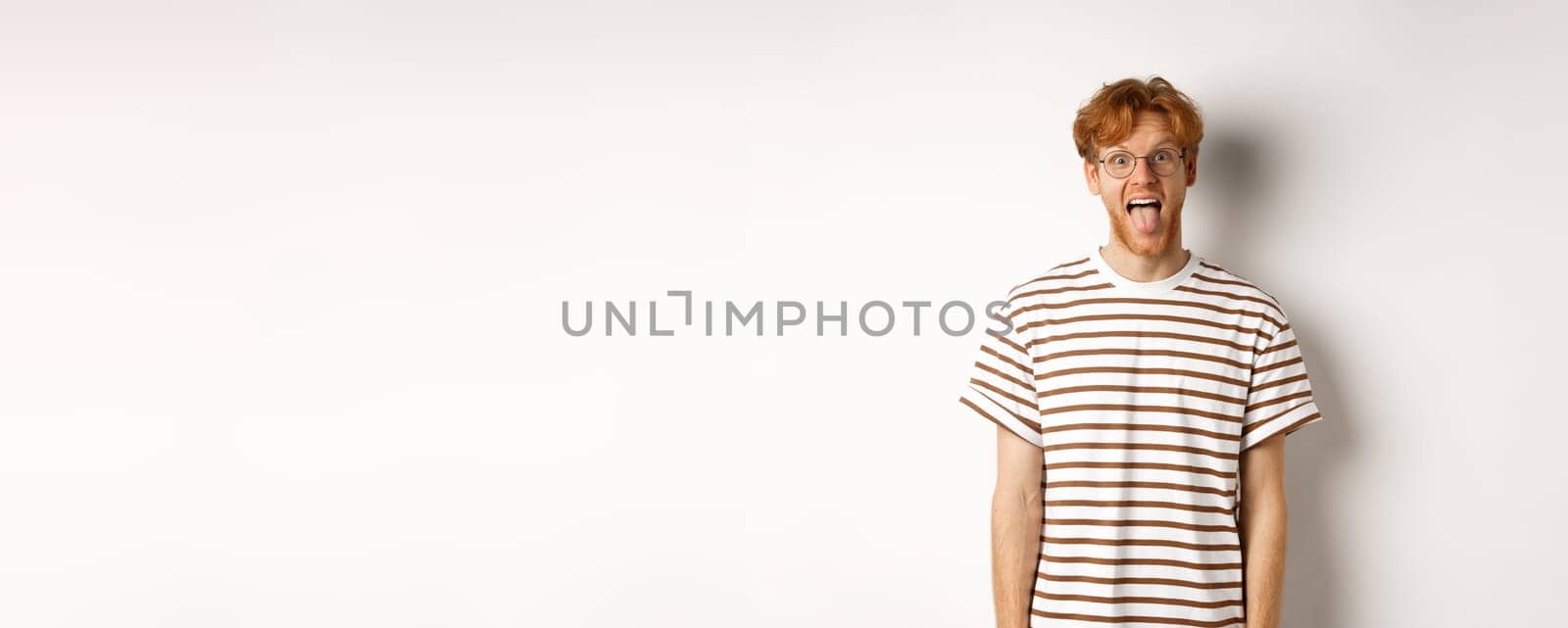 Funny young man with messy red hair and glasses showing tongue, staring at camera, standing over white background.