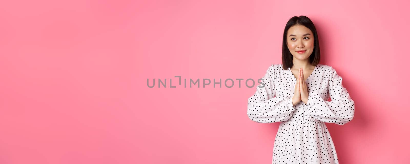Beautiful angelic asian woman smiling, holding hands in pray and looking left at copy space with innocent cute gaze, standing over pink background.