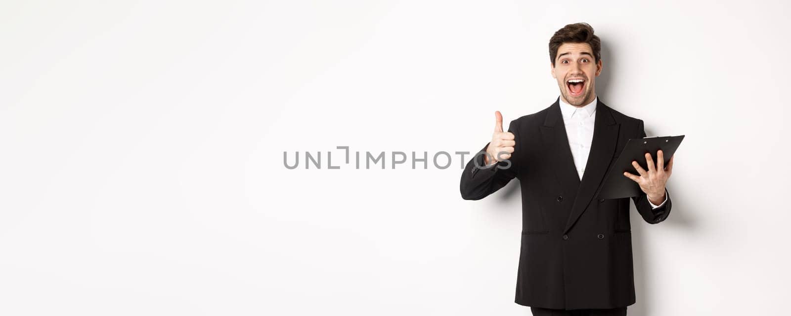 Portrait of confident businessman in black suit, holding clipboard with documents and showing thumb-up in approval, praise good job, standing against white background.