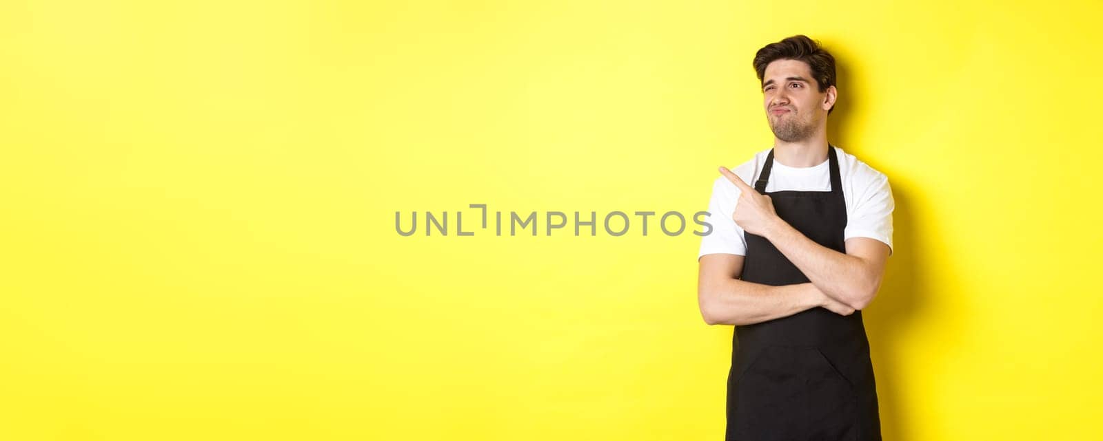 Skeptical male seller in black apron looking displeased, grimacing and pointing left at advertisement, standing over yellow background.