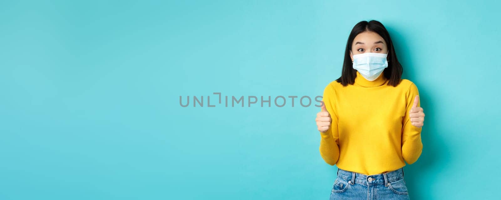 Covid-19, social distancing and pandemic concept. Excited asian woman showing thumbs up and looking impressed, praising good deal, wearing face mask, blue background.