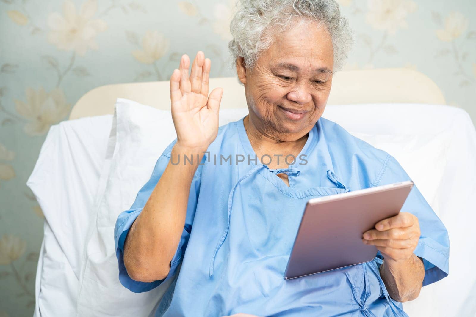 Asian senior woman patient holding in her hands digital tablet and reading emails while sitting on bed in hospital, healthy strong medical concept