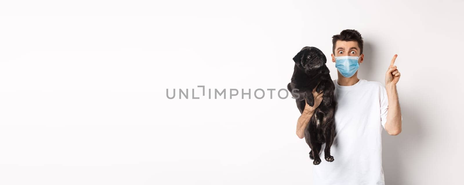 Covid-19, animals and quarantine concept. Young man in medical mask holding cute black pug, dog looking right and owner pointing at logo, white background by Benzoix