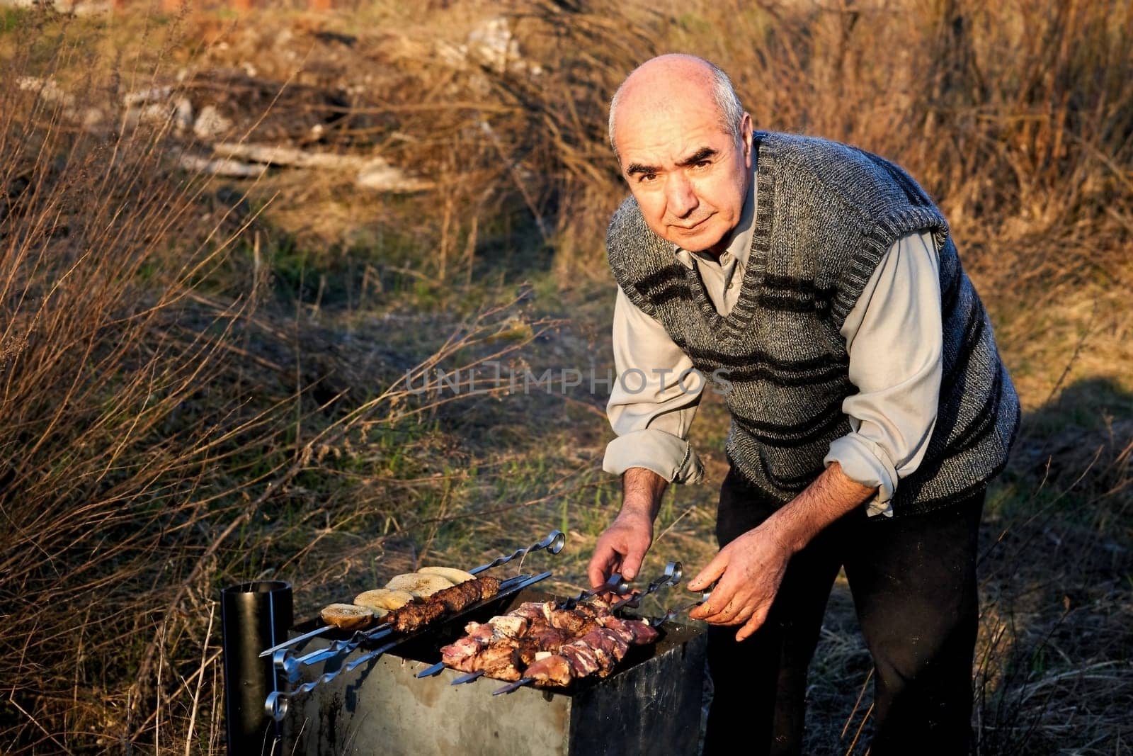 Elderly cheerful man pensioner cooking meat on the grill outdoors in nature by jovani68