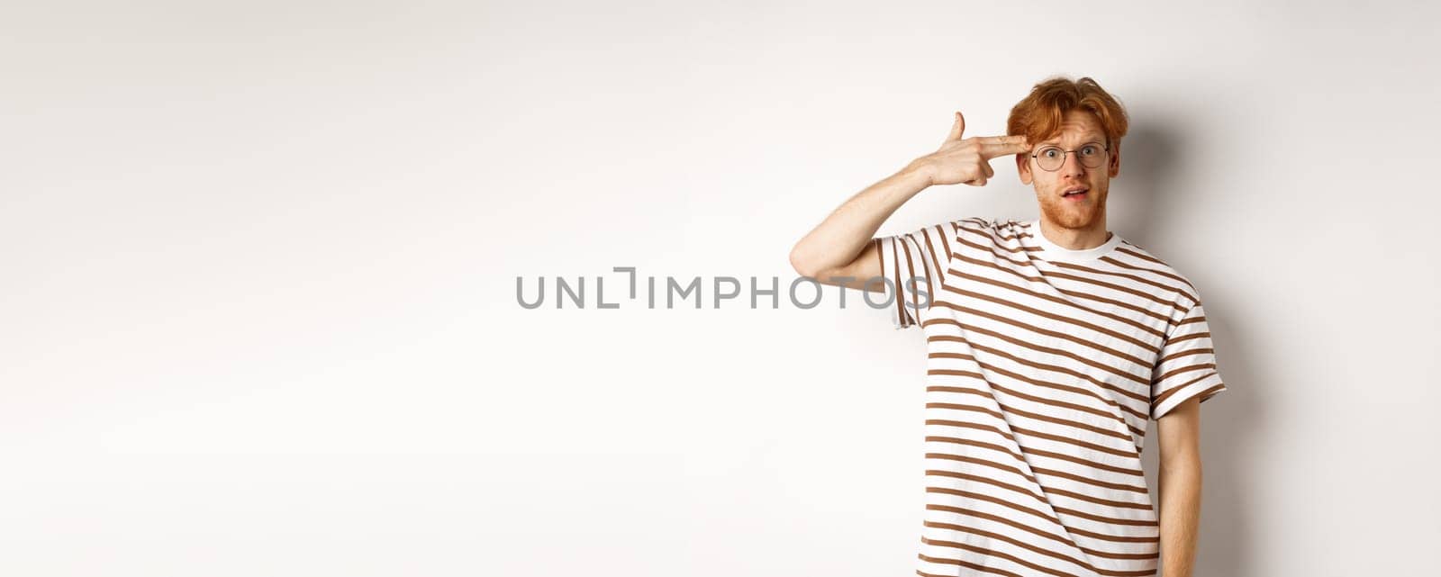 Annoyed teenage redhead guy making finger gun near head, staring bothered and irritated at camera, white background.