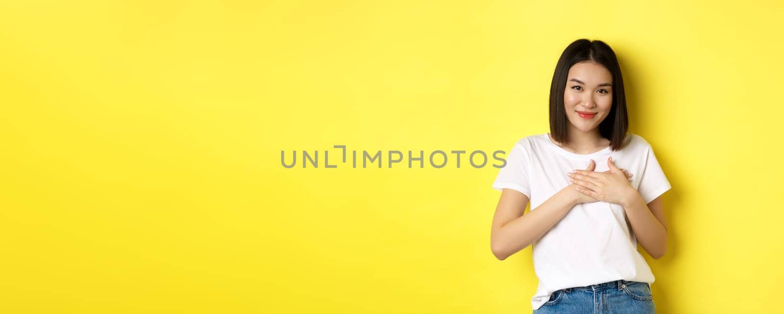 Beauty and fashion concept. Heartfelt asian girl holding hands on heart and smiling touched, thanking you, standing over yellow background.