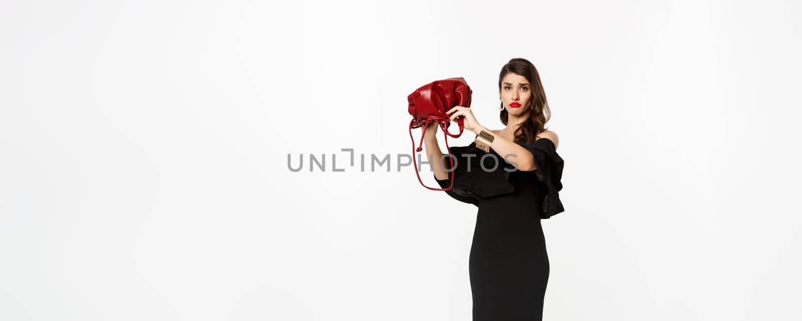 Beauty and fashion concept. Full length of sad young woman in black dress and high heels showing empty purse, sulking disappointed, standing over white background.
