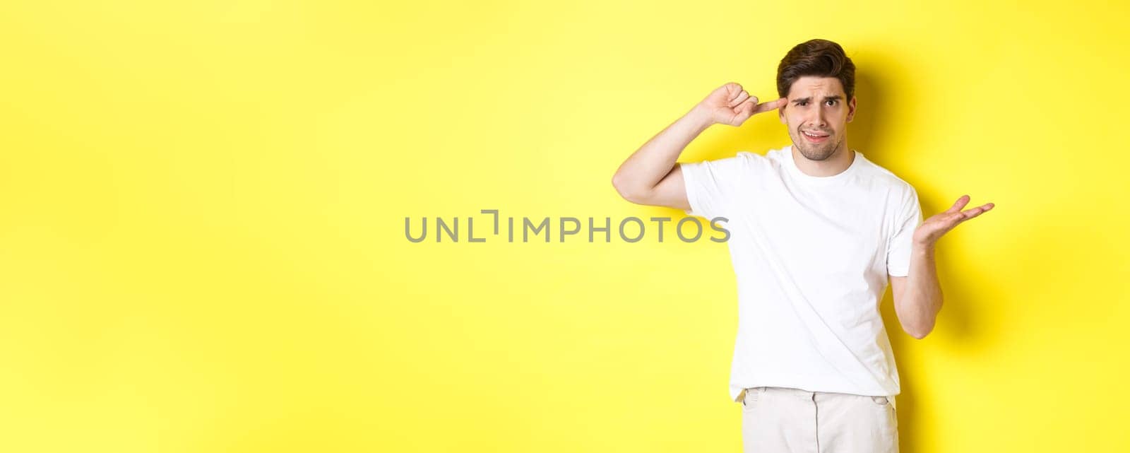 Confused and angry man pointing at head, scolding person for acting stupid, show crazy sign, standing over yellow background.