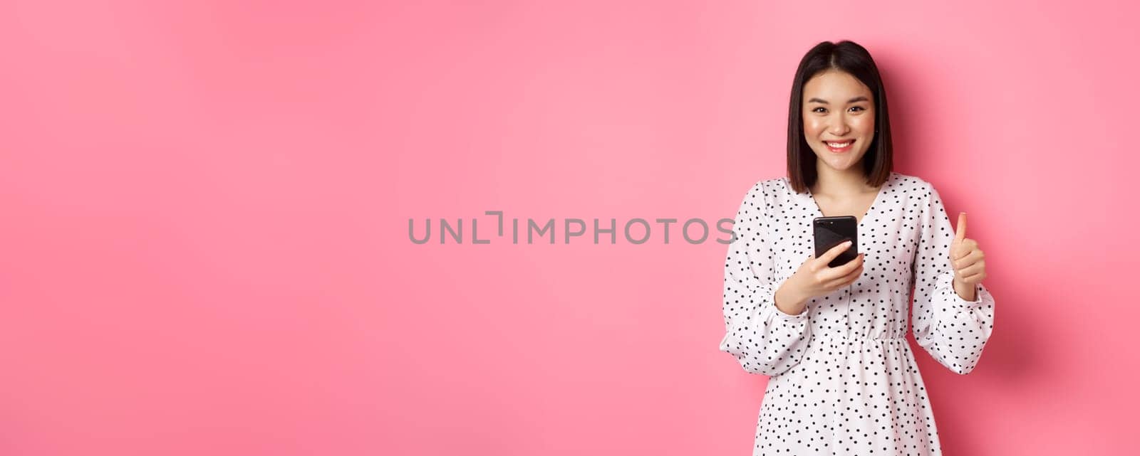 Online shopping and beauty concept. Satisfied asian female customer showing thumbs-up, making purchase in internet on smartphone, standing over pink background.