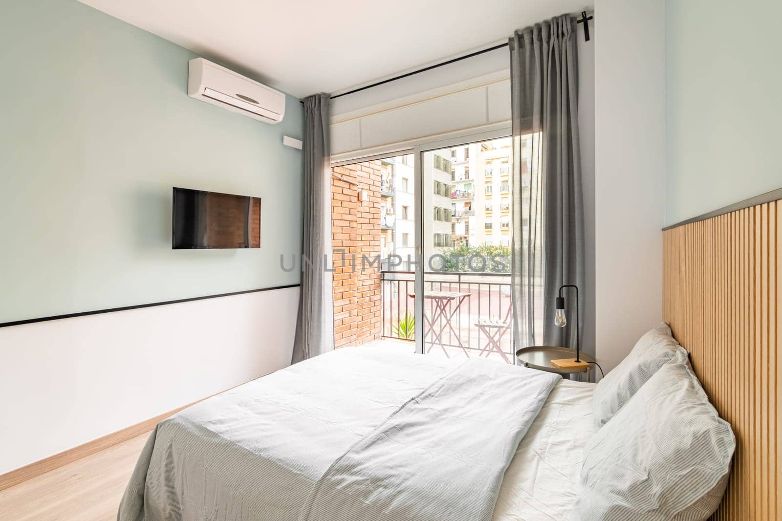 Small but pleasant bedroom with a double bed with white linens, TV and an open terrace overlooking the courtyard of a residential complex or tourist hotel on a sunny summer day.