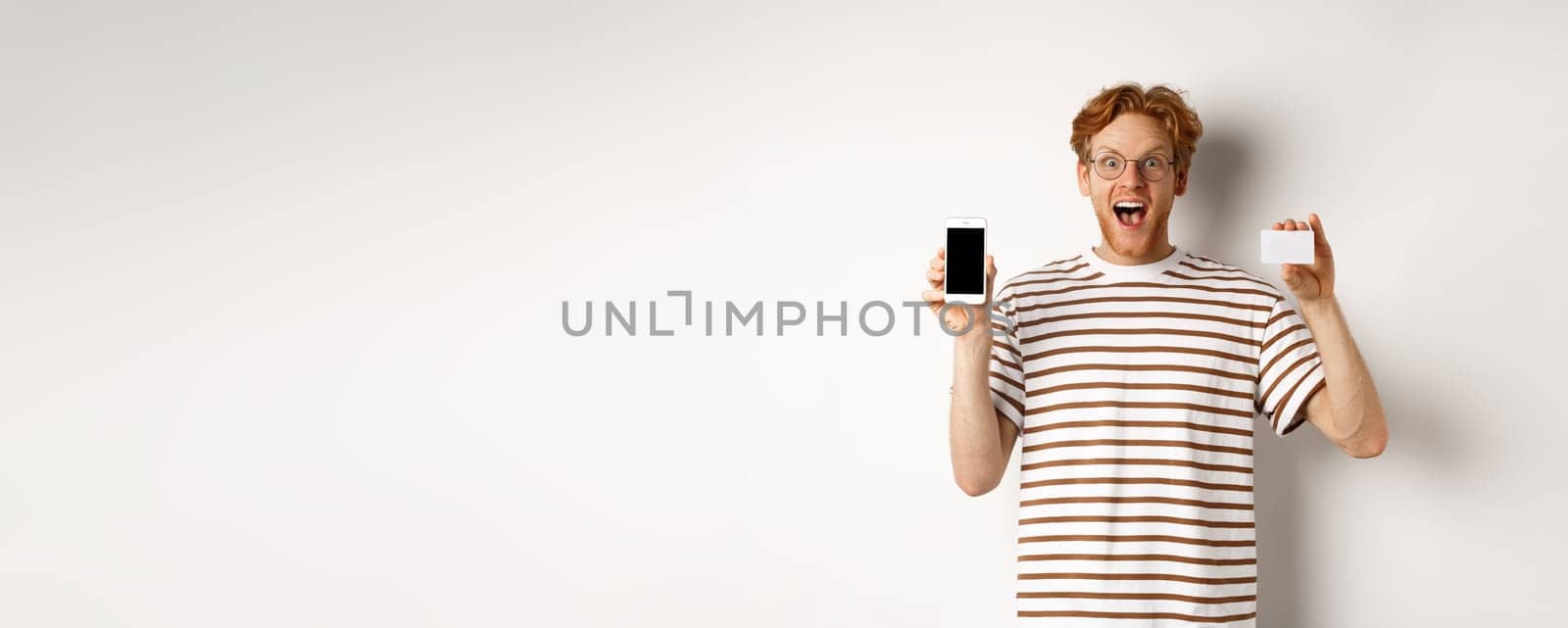 Shopping and finance concept. Young man showing blank mobile screen and plastic credit card, smiling at camera, white background by Benzoix