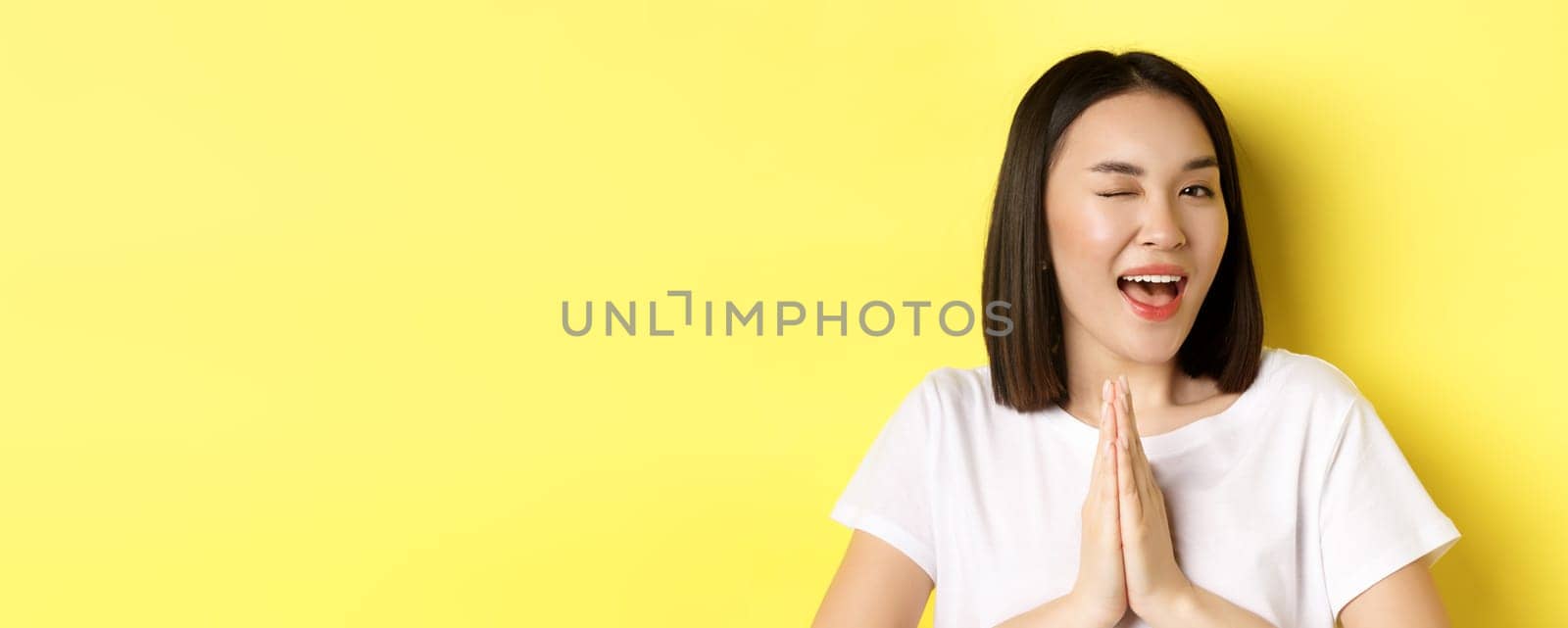 Close of cheeky young asian woman holding hands in namaste, thank you gesture, winking at camera coquettish, feeling lucky, standing over yellow background.