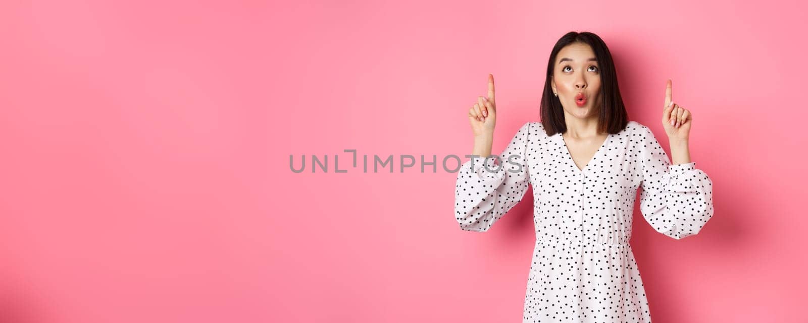 Cute korean girl in beautiful dress saying wow, looking and pointing fingers up, intrigued in promo offer, standing over pink background.