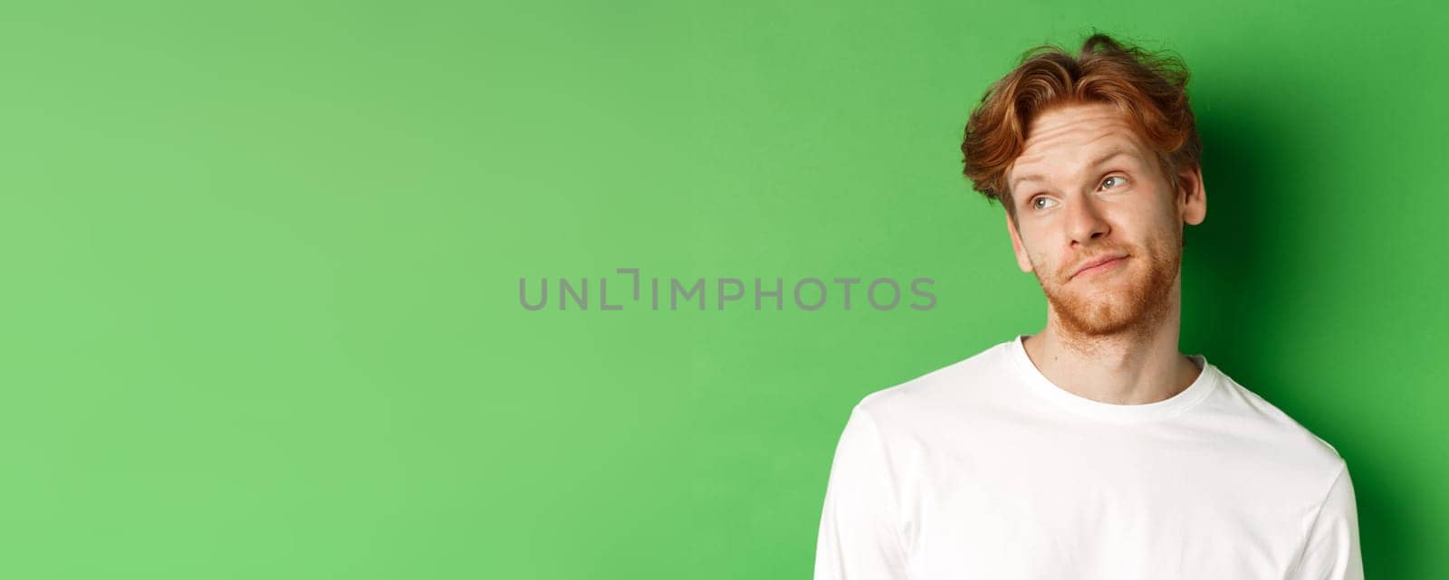Smiling redhead man with messy haircut and beard tilt head, looking left with pleased face, standing over green background by Benzoix