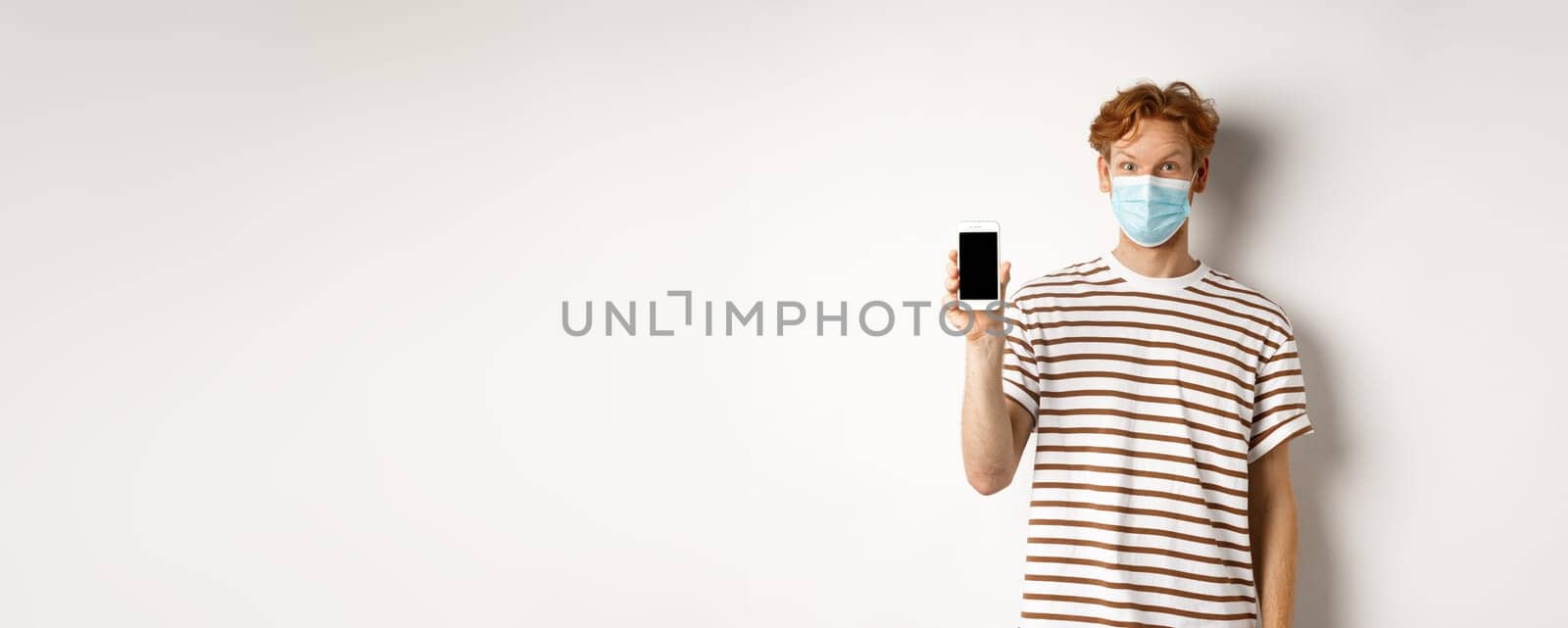 Covid-19, online doctor concept. Smiling redhead man in face mask showing smartphone screen, standing over white background by Benzoix