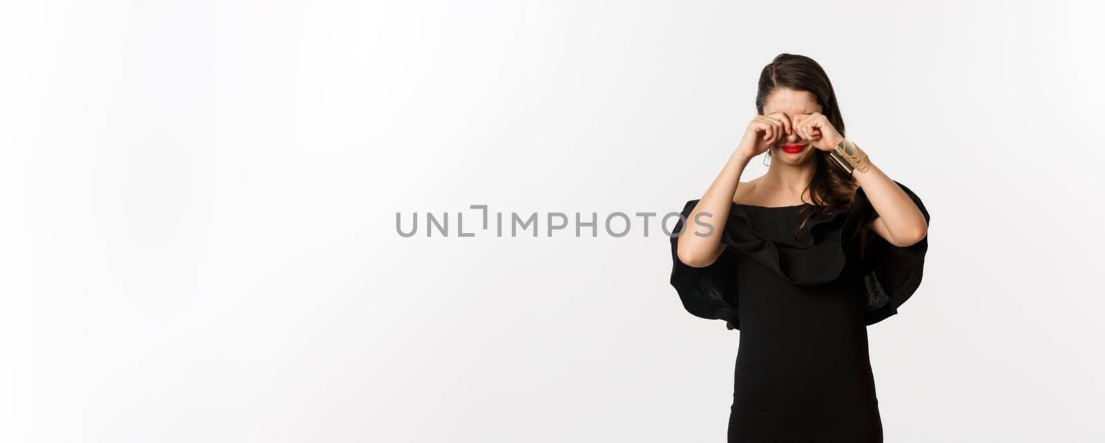 Fashion and beauty. Sad and gloomy woman in black dress crying, rubbing eyes and sulking, standing over white background.