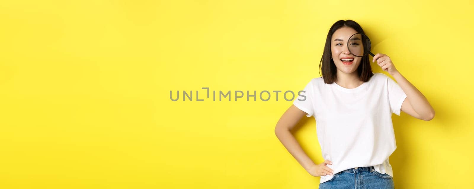 Cheerful asian girl searching for you, looking through magnifying glass and smiling, found something interesting, standing over yellow background by Benzoix
