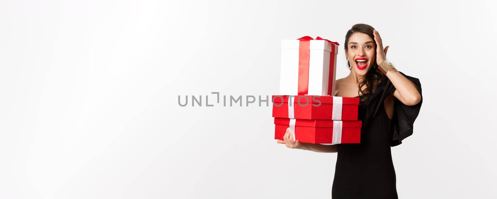 Celebration and christmas holidays concept. Excited and happy woman receive gifts, holding xmas presents and rejoicing, standing in black dress over white background.