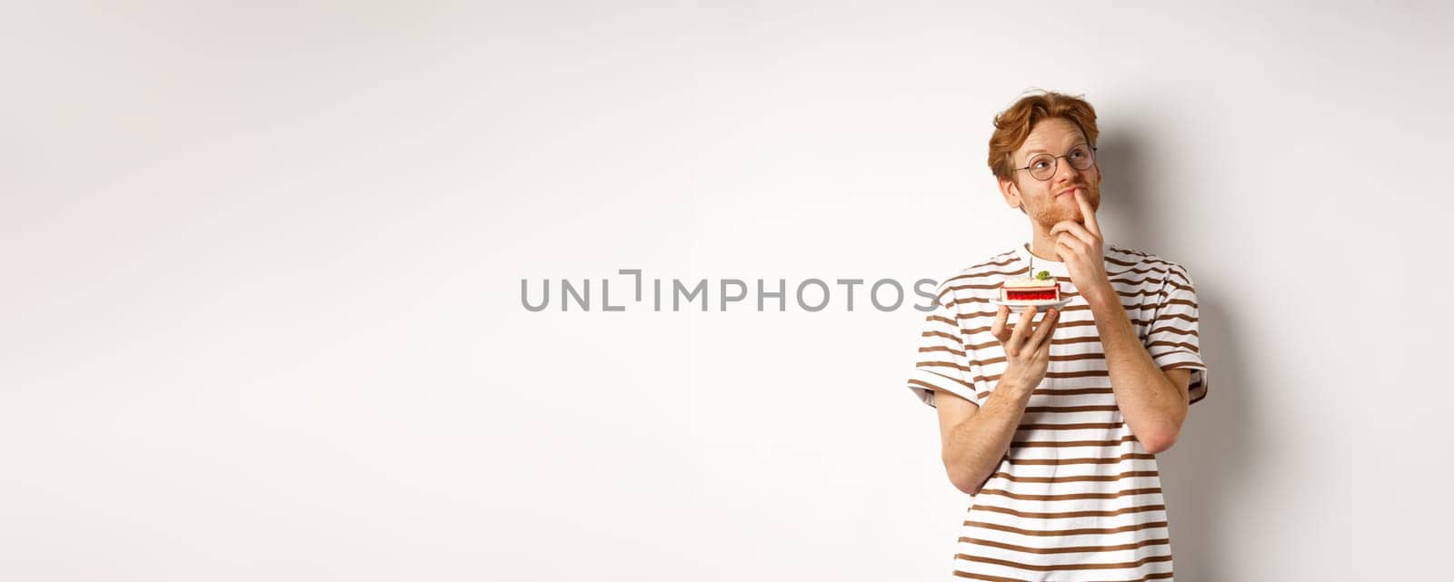 Holidays and celebration concept. Happy young man with red hair and glasses having birthday, holding cake with candle and thinking of b-day wish, standing over white background.