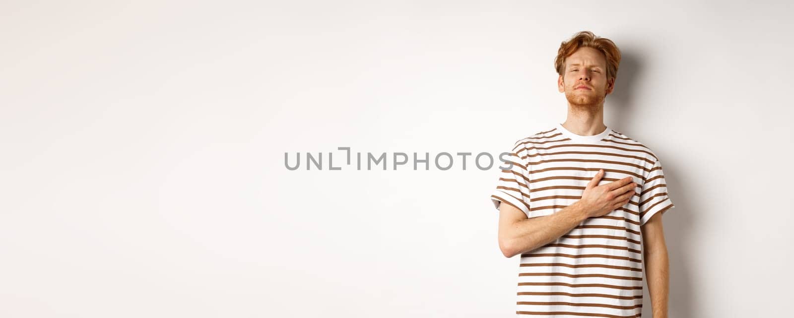 Young bearded redhead man holding hand on heart and looking patriotic, standing during national anthem, standing over white background by Benzoix