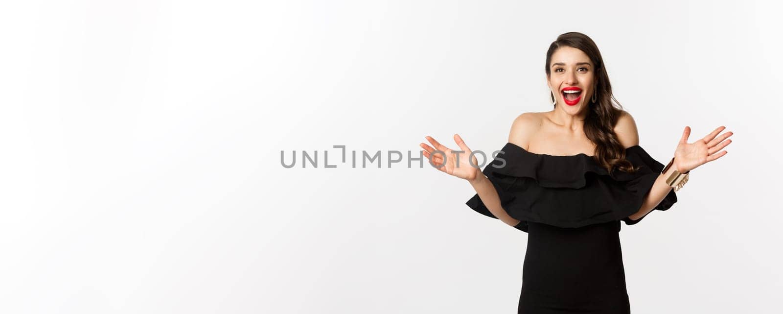 Beauty and fashion concept. Image of surprised and happy young woman in party dress reacting to good news, raising hands up and smiling amazed, white background by Benzoix
