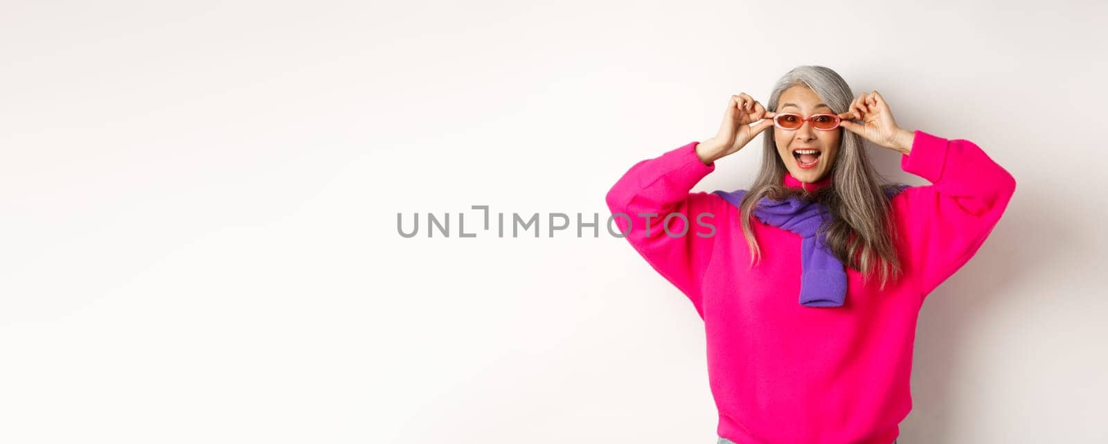 Fashionable asian senior woman trying new sunglasses, looking amazed and cheerful at camera, standing in trendy sweater against white background.