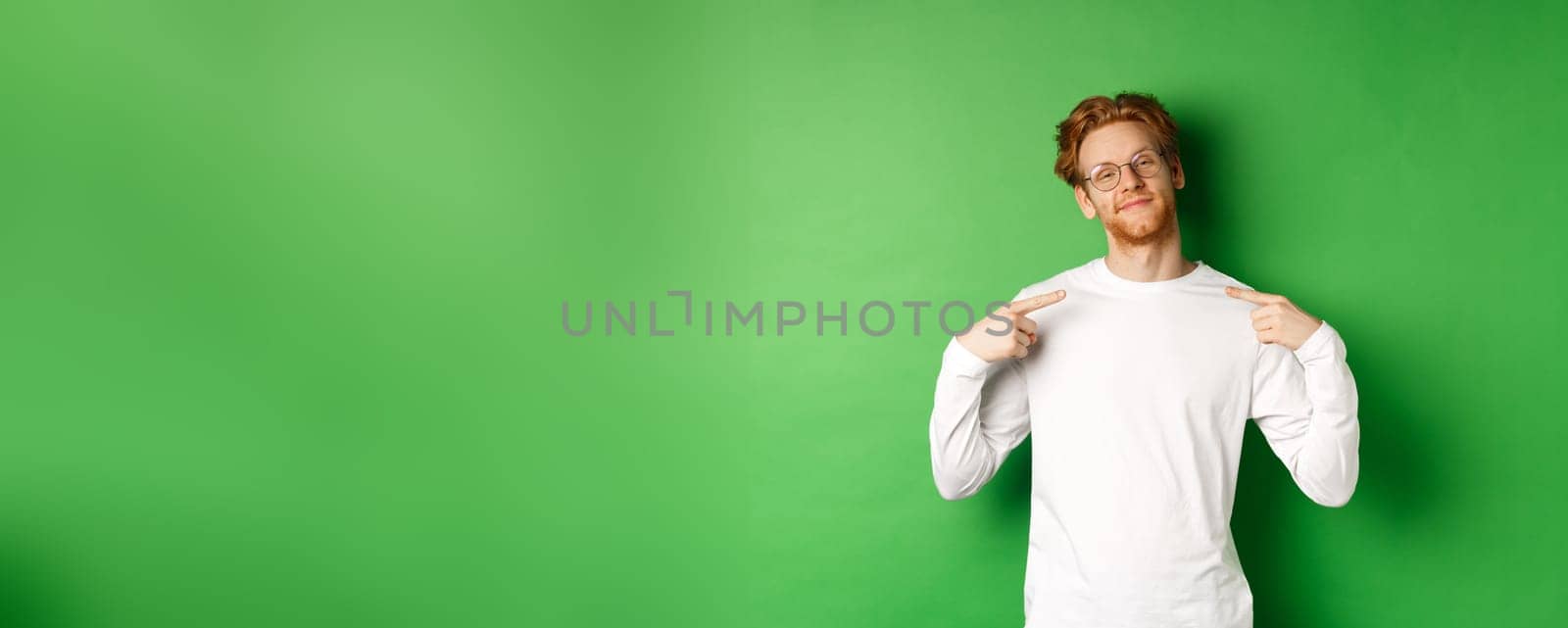 Confident and smug redhead man in glasses smiling, pointing at himself self-assured, standing over green background. Copy space