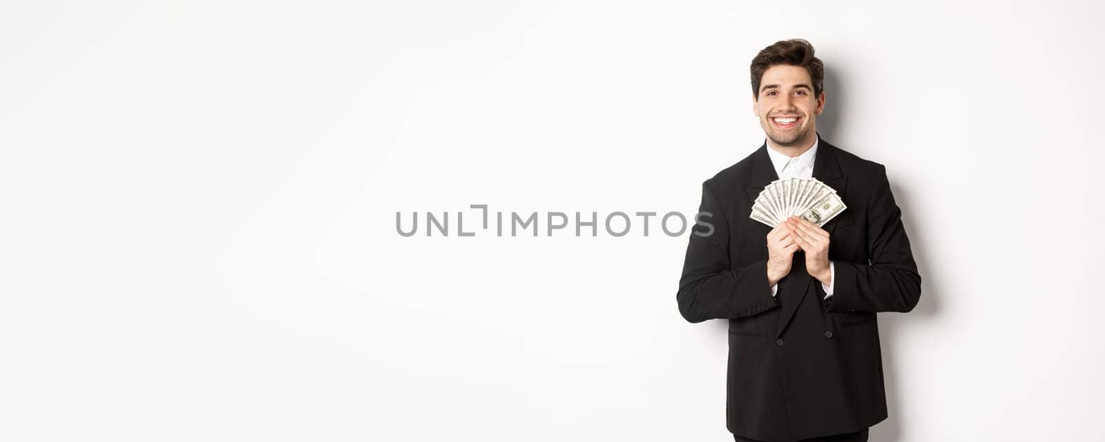 Portrait of happy and pleased handsome man in suit, hugging money and looking satisfied, standing over white background.