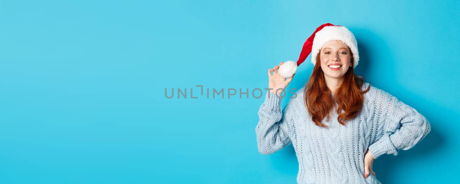 Winter holidays and Christmas Eve concept. Cheerful redhead teenage girl wearing Santa hat and smiling satisfied, standing in sweater against blue background.