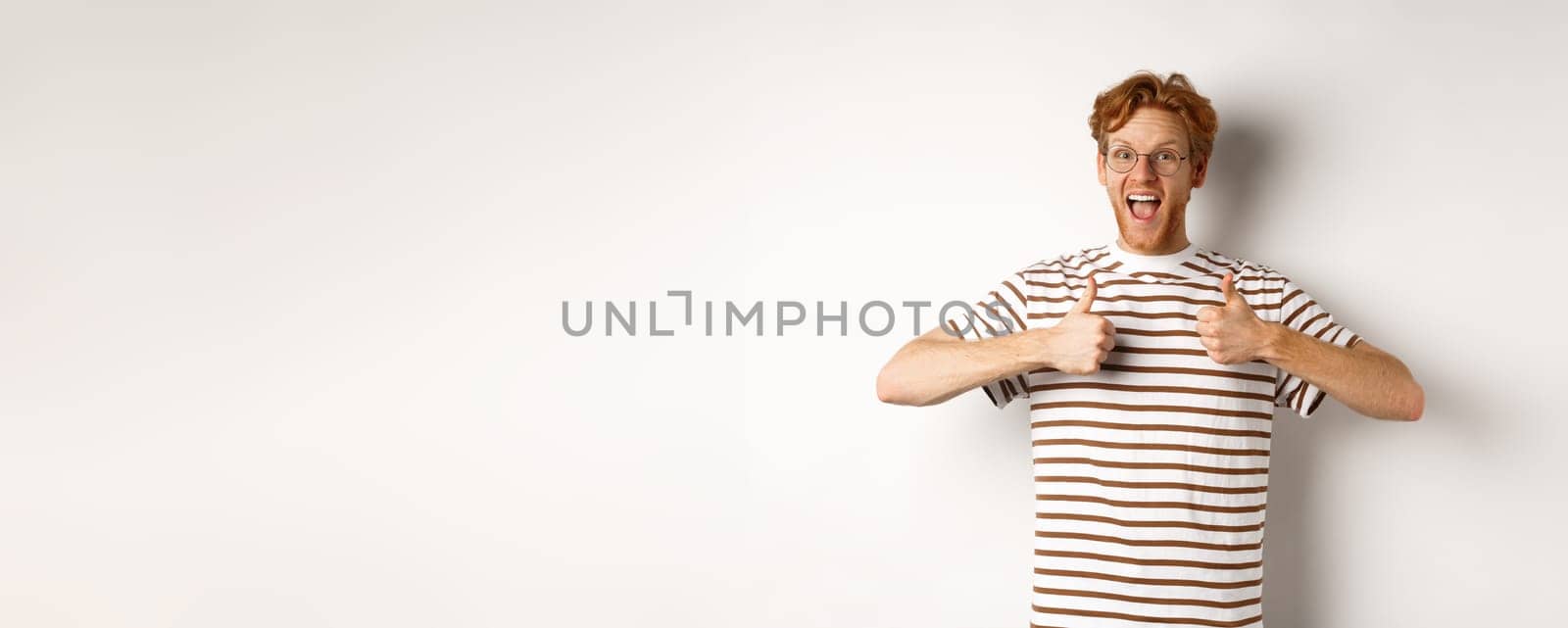 Young amazed man in red hair checking out something awesome, saying yes and showing thumbs-up, standing over white background.