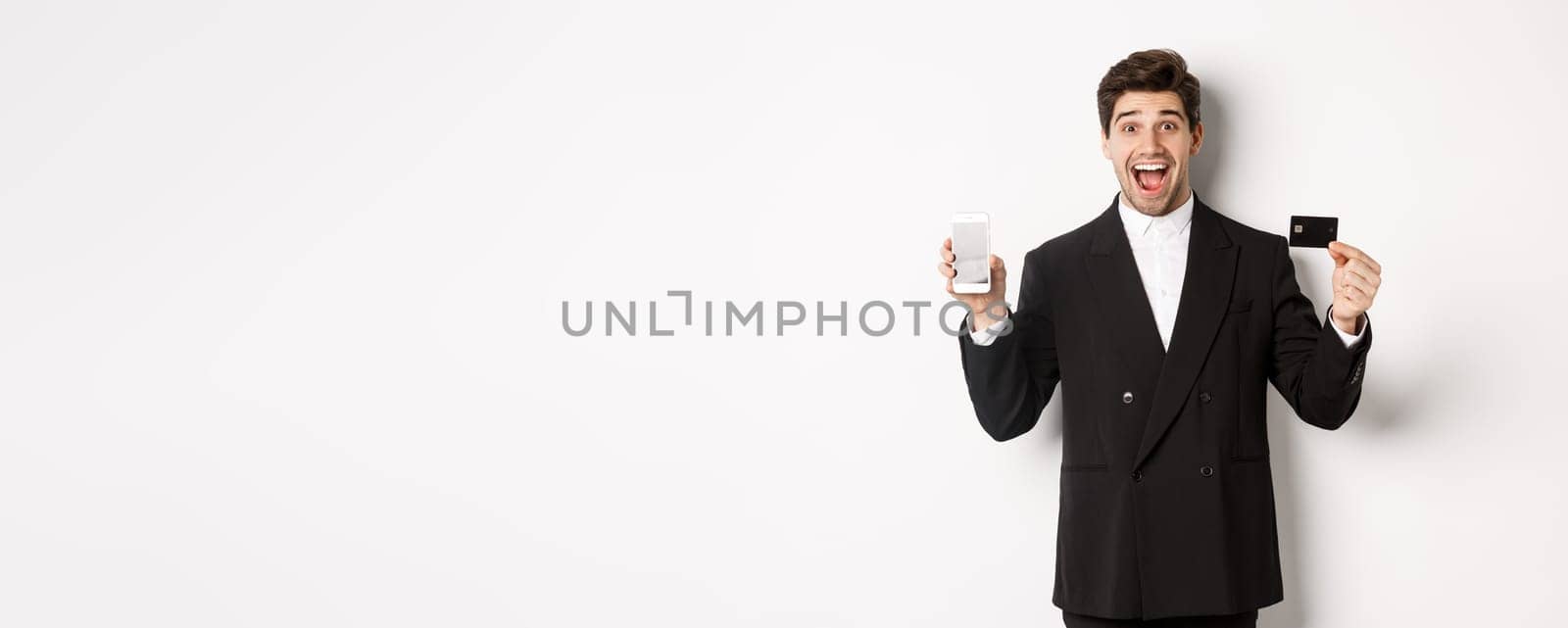 Image of handsome businessman in black suit, looking excited and showing credit card with mobile phone screen, standing against white background by Benzoix