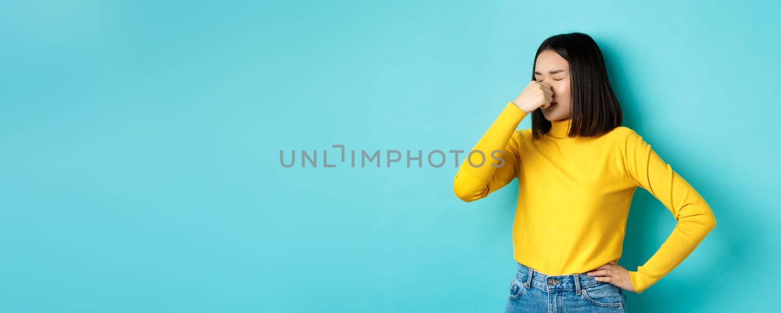 Image of asian stylish girl shut her nose and grimacing from aversion, something smell bad, standing disgusted against blue background.