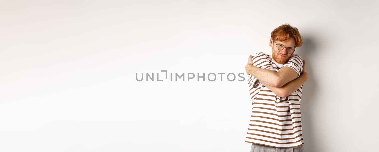 Reluctant young man with messy red hair unwilling do something, hugging himself and grimacing, standing over white background.