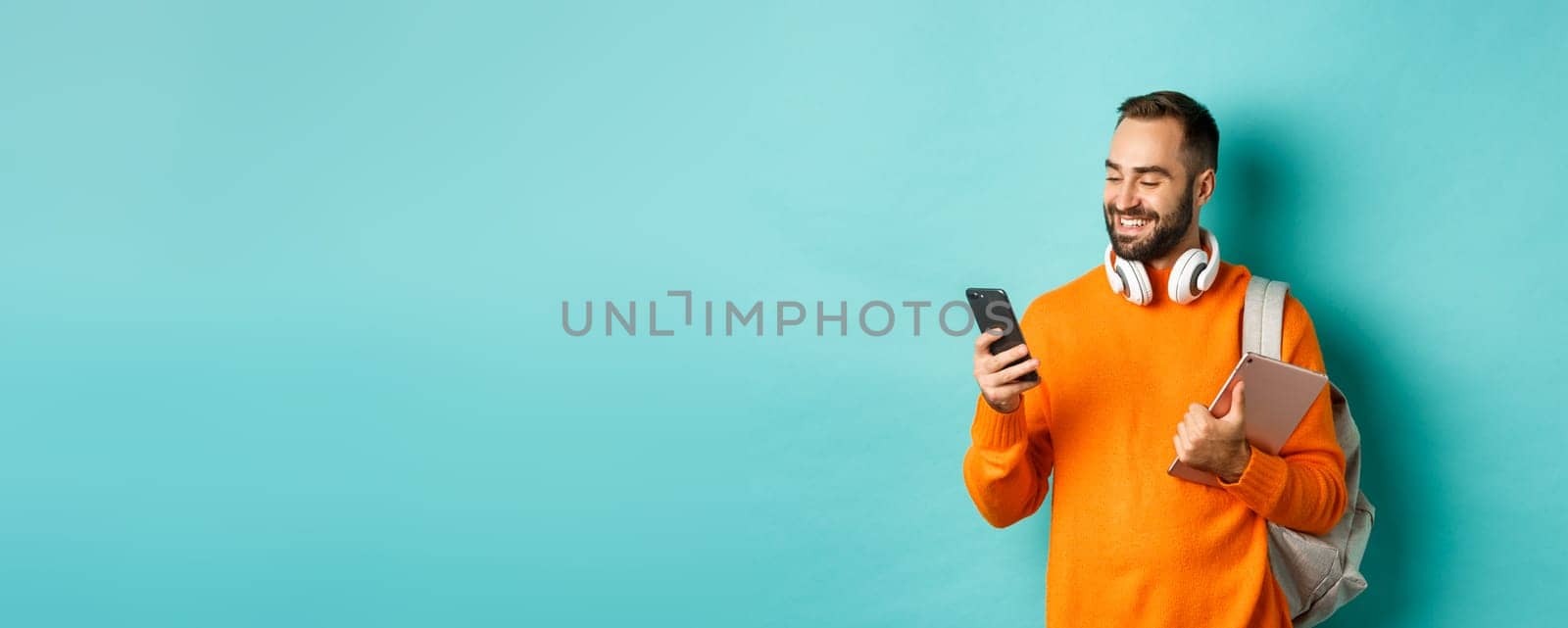 Handsome man student with headphones and backpack, holding digital tablet, reading message on mobile phone, standing against light blue background.