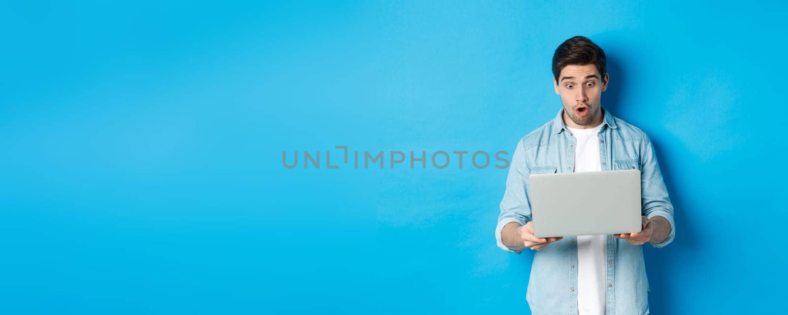 Impressed caucasian guy looking at laptop screen with amazement, checking out promo in internet, standing against blue background by Benzoix