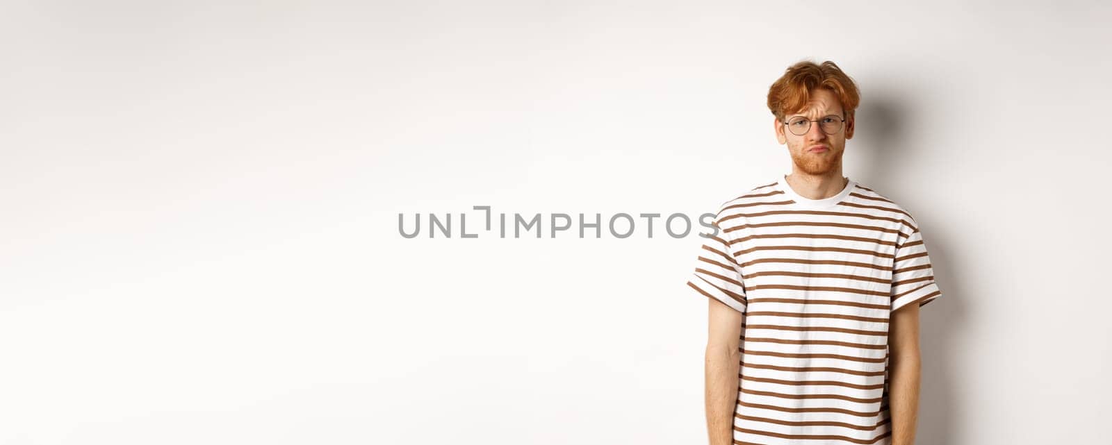 Sad and gloomy redhead male student pouting, frowning and looking distressed, standing over white background.
