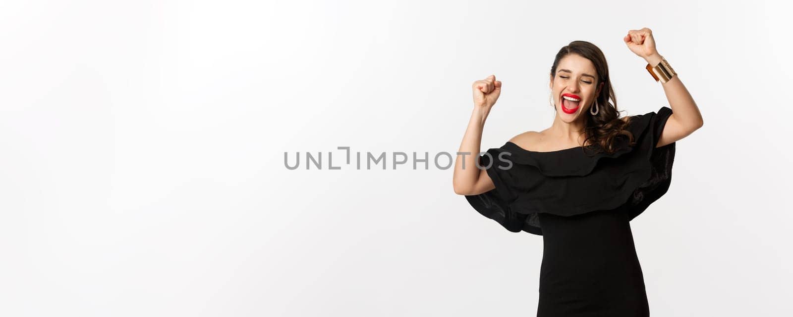 Fashion and beauty. Successful pretty woman in black dress celebrating, rejoicing of winning, triumphing over white background.