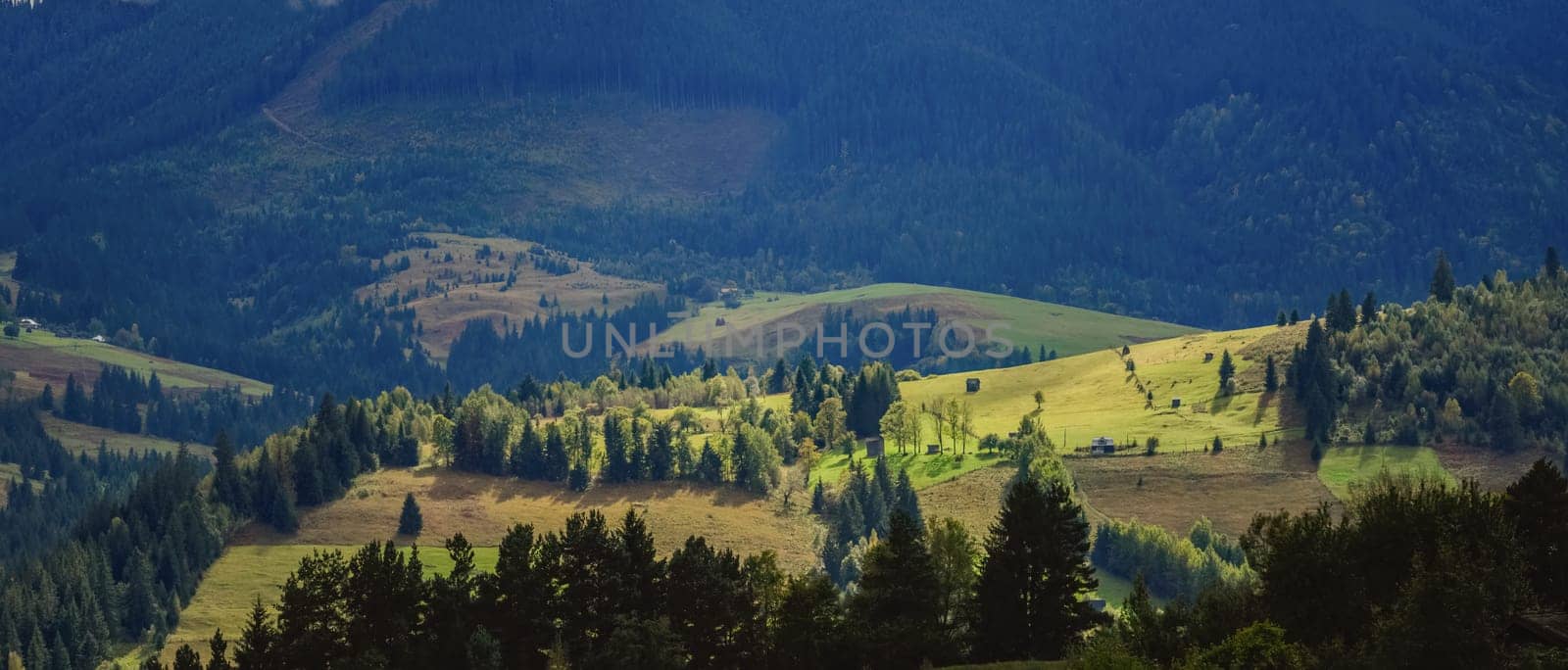 Wonderful mountain land with forest and small houses in sunlight. Scenic view to vast expanses of rolling hills and great mountain ridge on horizon. Immense distances of wavy relief in evening light. by Renisons
