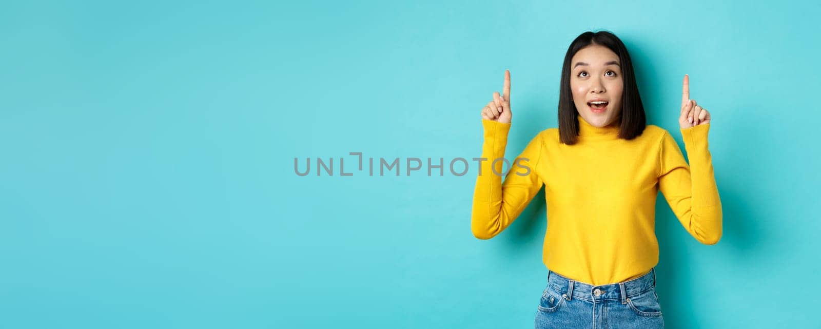 Shopping concept. Dreamy korean woman looking with desire at promo offer, pointing fingers up and staring at advertisement amazed, standing over blue background.