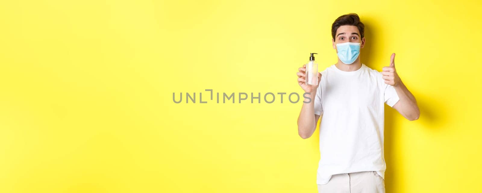Concept of covid-19, quarantine and lifestyle. Satisfied young man in medical mask showing good hand sanitizer, thumbs up and recommending antiseptic, yellow background.