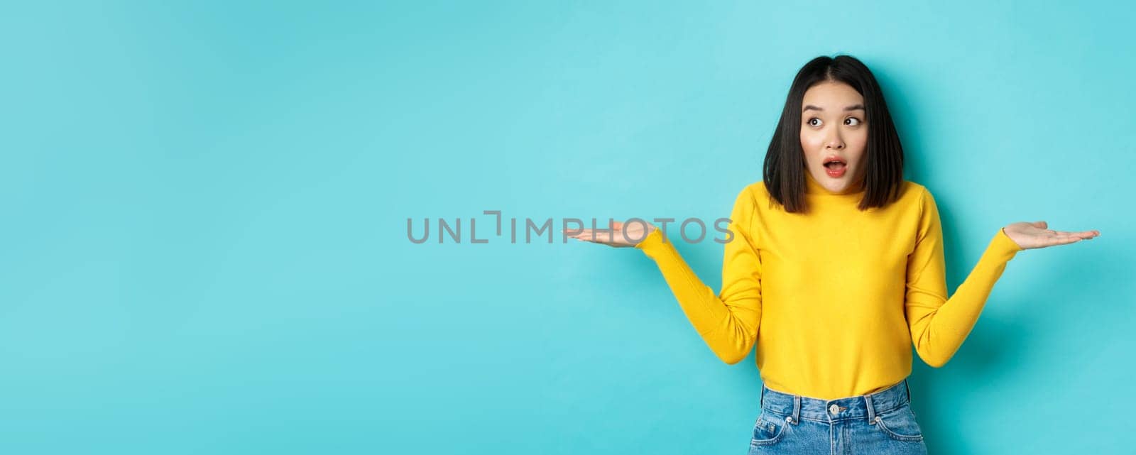Confused and shocked asian woman dont understand, shrugging with spread hands, gasping and looking left, know nothing, standing over blue background by Benzoix