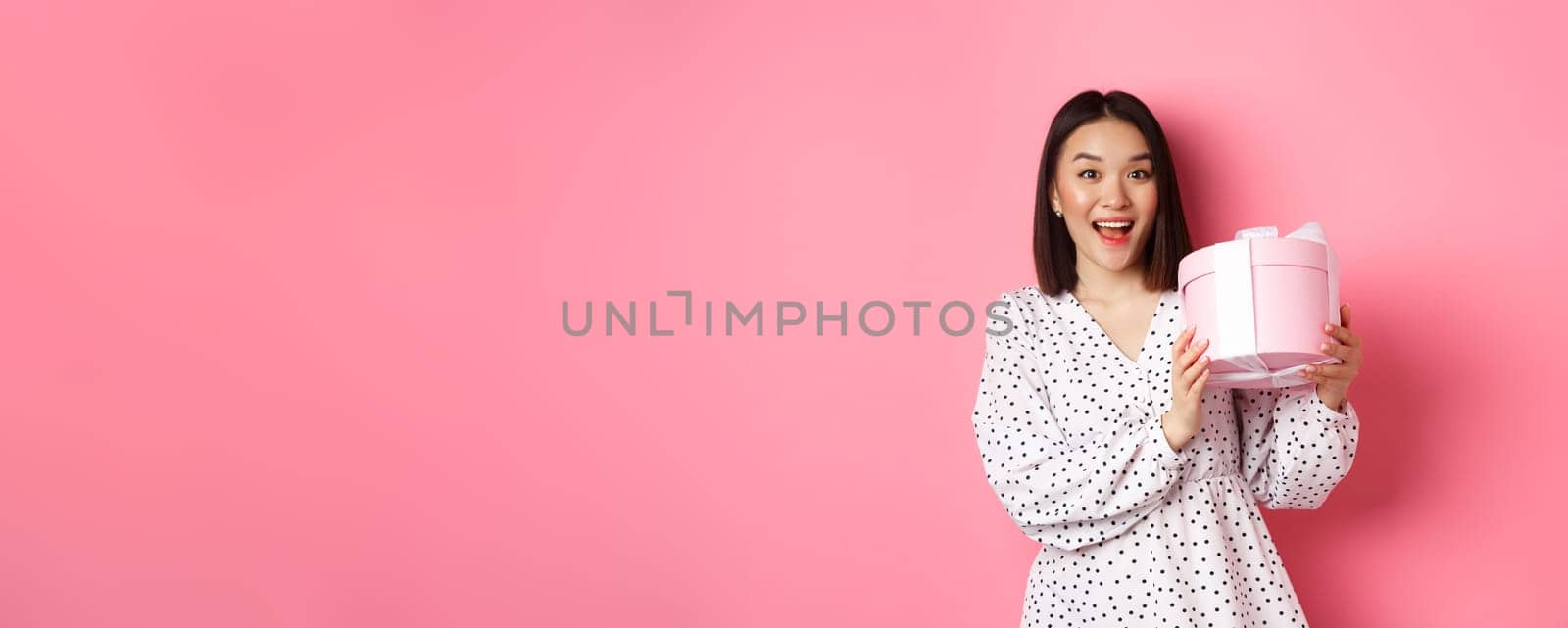 Valentines day. Happy asian woman receiving gift in cute box, smiling excited and thankful, standing in dress against pink background by Benzoix