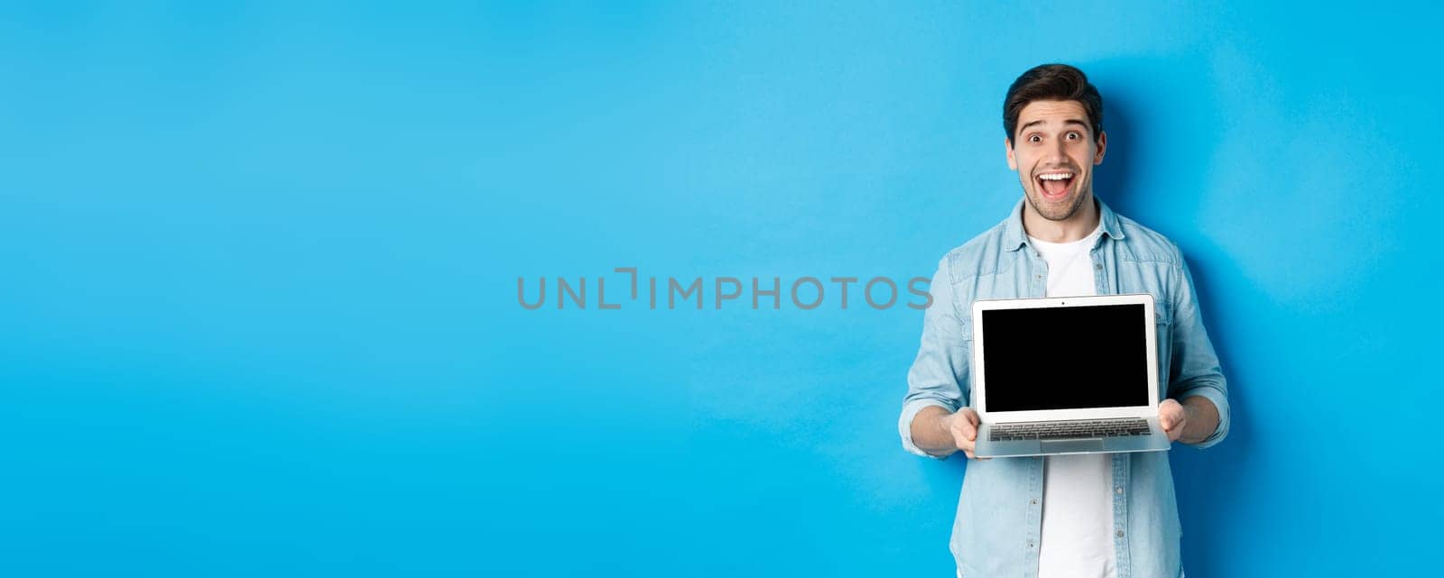 Cheerful smiling man making presentation, showing laptop screen and looking happy, standing over blue background.