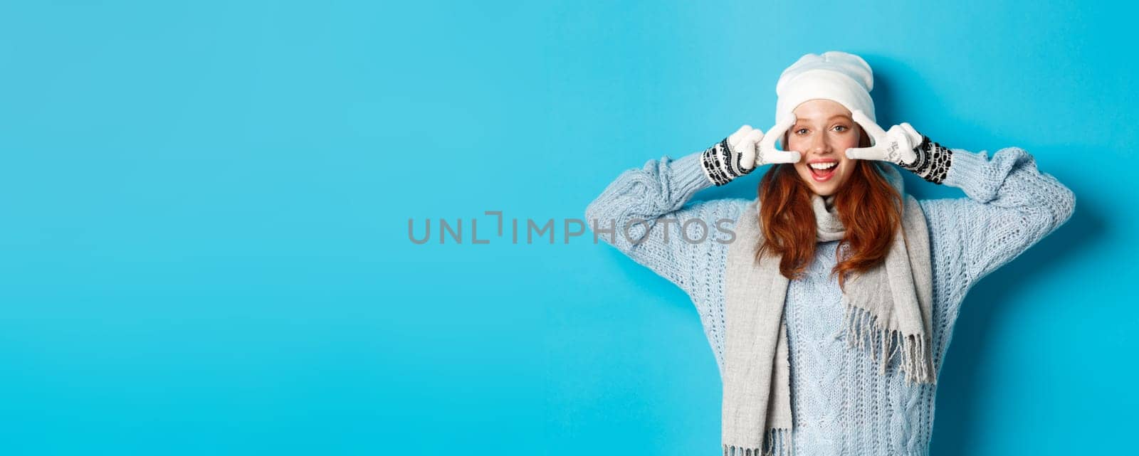 Winter and holidays concept. Cute redhead teen girl in beania, gloves and sweater showing peace sign, looking left at camera and wishing merry christmas, standing against blue background by Benzoix