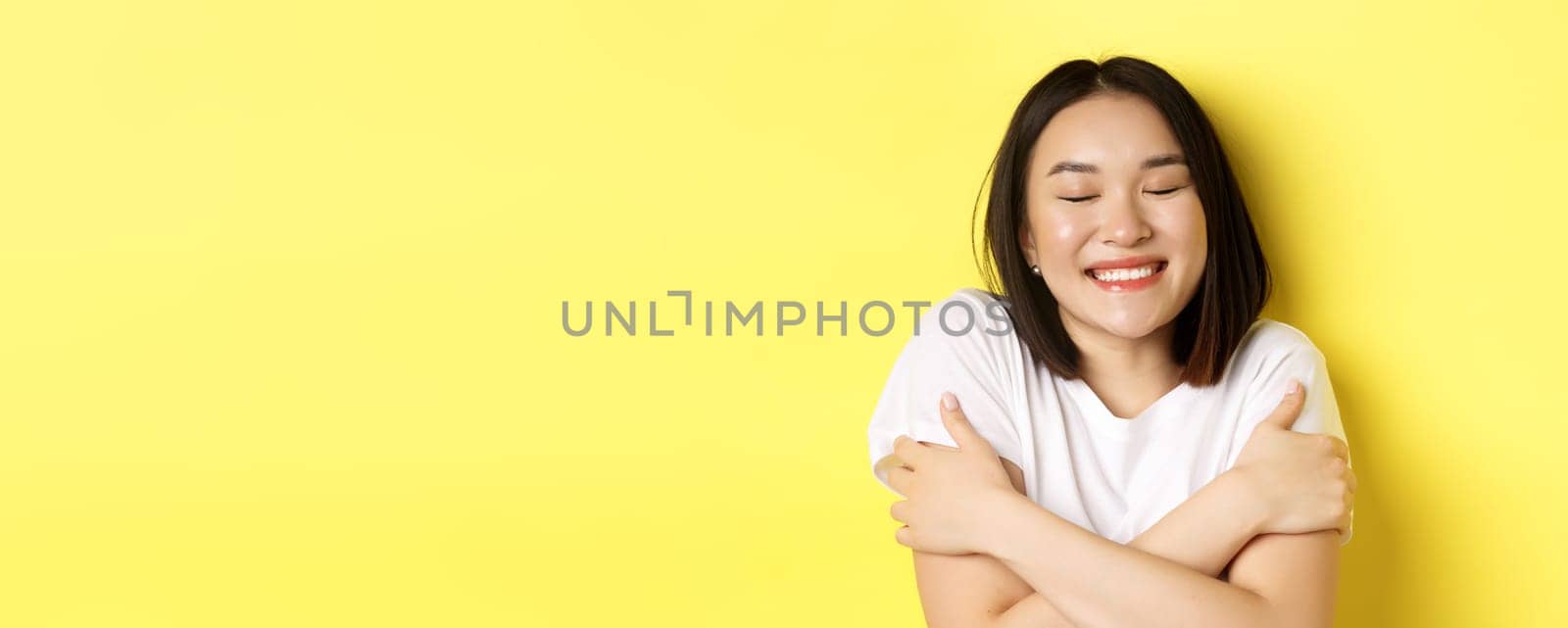 Close up of romantic asian girl hugging herself and dreaming, close eyes and smile while imaging something tender, standing over yellow background.