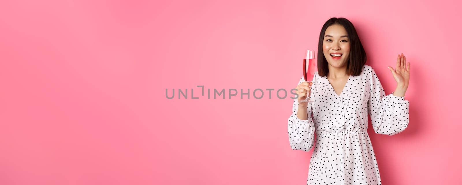 Celebration. Beautiful asian woman drinking champagne and smiling, standing in dress over pink background by Benzoix