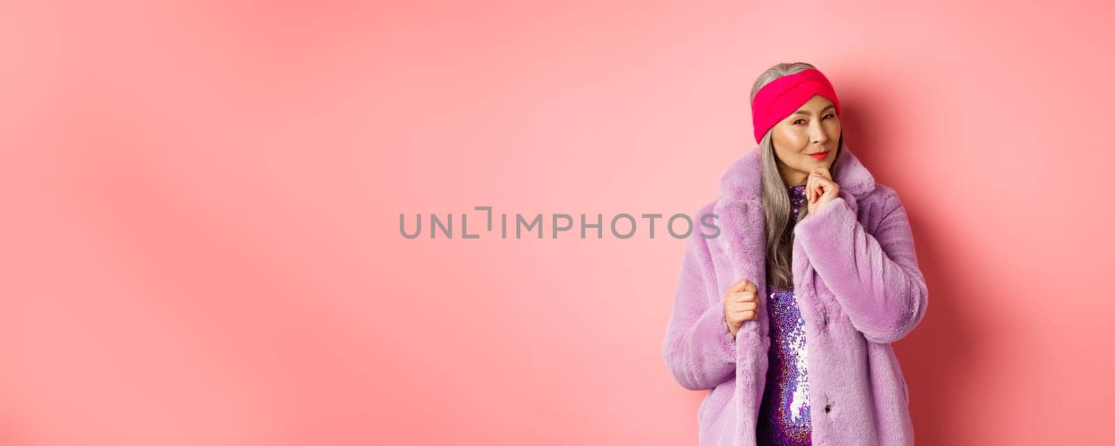 Fashion and shopping concept. Stylish old asian lady in purple fake-fur coat looking intrigued, interested with promotion, smiling and thinking, pink background.