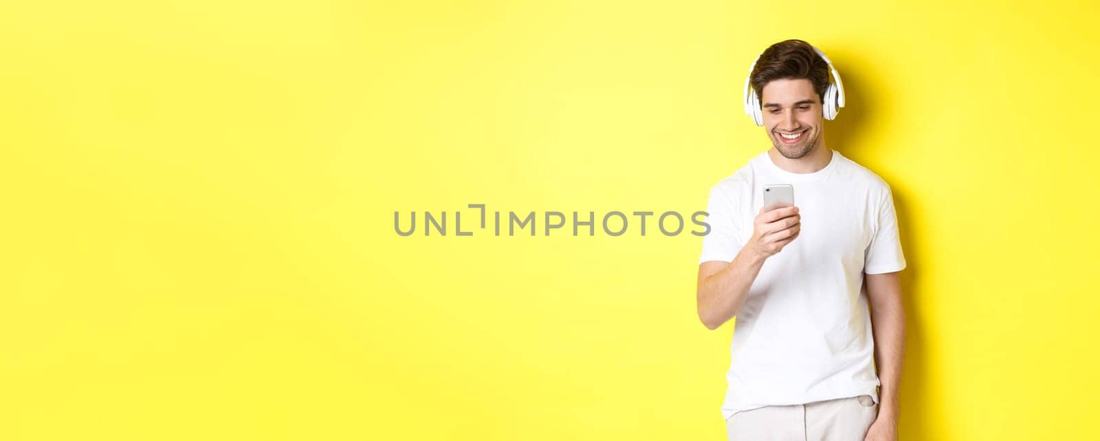 Young man in headphones reading message on smartphone, smiling, listening music in earphones and picking song in app, standing over yellow background.