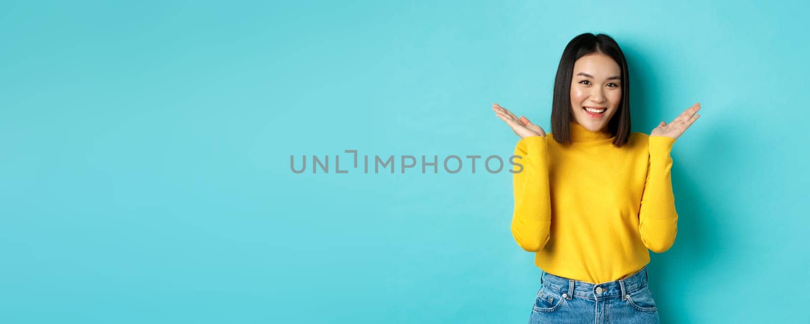 Beauty and fashion concept. Attractive japanese girl raise hands up and demonstrate something, smiling happy and looking at camera, showing promo, blue background by Benzoix